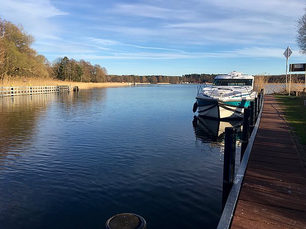 Zu sehen ist ein Steg mit einem Kunststoff-Hausboot an einem Kanal, im Hintergrund ist ein großer See zu sehen, dessen Schilfrand noch winterlich-braun ist.